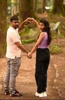 A couple stands in the woods, their hands joined to form a heart shape, framed by trees and soft, natural light.