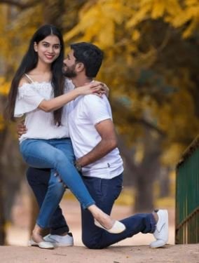A couple smiles and poses together for a photo in a lush green park, surrounded by trees and sunlight.