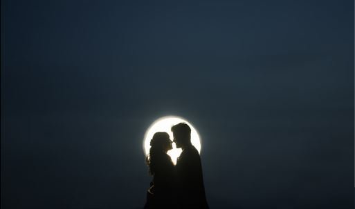 A couple shares a romantic kiss beneath a glowing moon, surrounded by a serene night sky filled with stars.